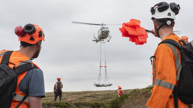 A helicopter flying over moorland  with large white bags of hanging from wires from underneath it. 4 people wearing hard hats and orange hi-vis jackets are  helping it to deliver materials. One of them has a handful of orange flags